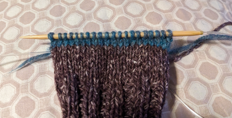 Partially knit sock with three rows of heel stitches on one side in blue waste yarn