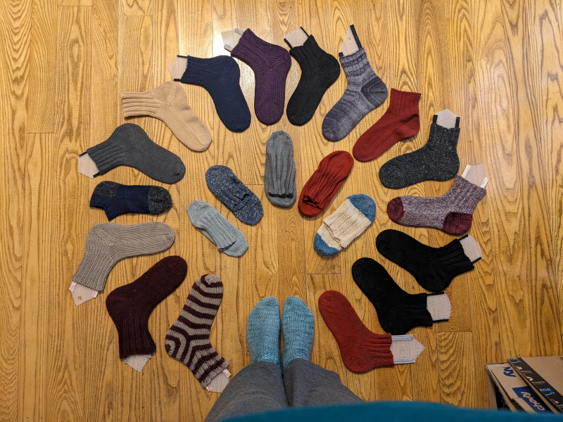 A circle of 16 hand-knit socks on sock blocks with my feet clad in another pair of hand knit socks completing the circle, in the center are 5 additional socks 